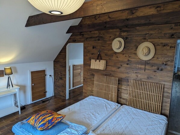 A bedroom with a sloped ceiling, wooden wall paneling, a bed with two pillows, a bedside table with a lamp, and a wardrobe.