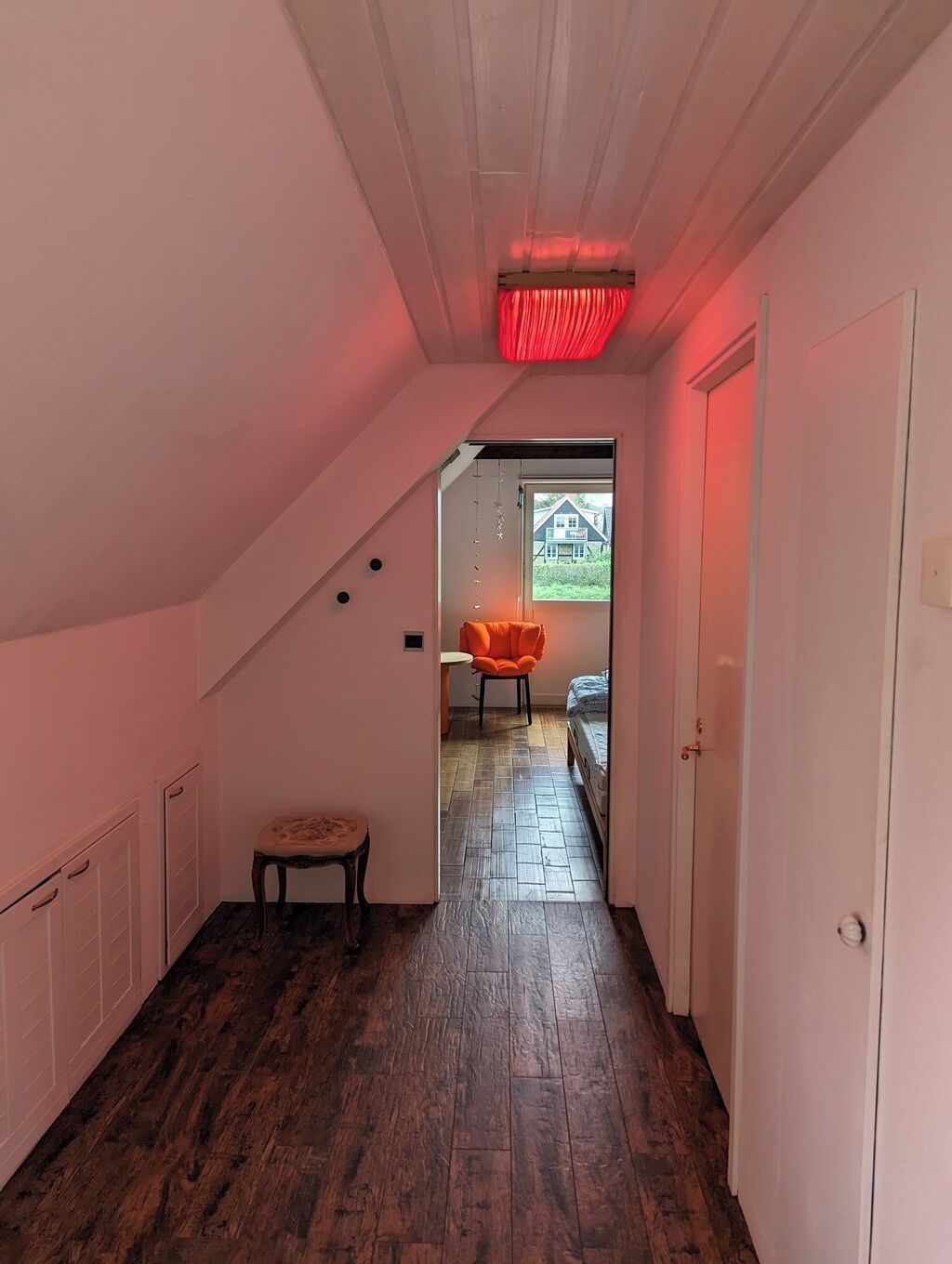 Een smalle gang met houten vloer, witte muren en een schuin plafond. Een feloranje stoel staat bij een deur die leidt naar een kamer met een bed.