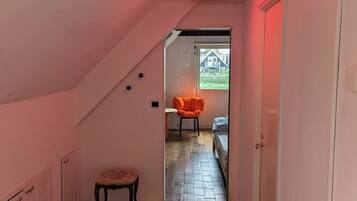 Een smalle gang met houten vloer, witte muren en een schuin plafond. Een feloranje stoel staat bij een deur die leidt naar een kamer met een bed.