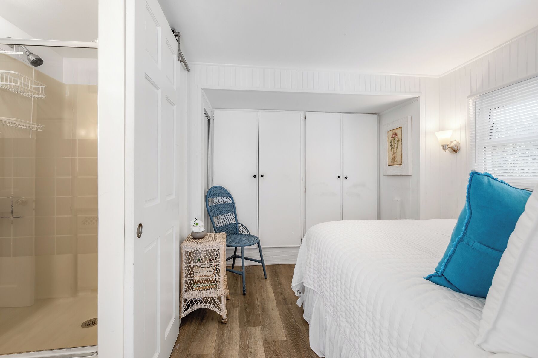 A bedroom with a bed, a chair, a small table, a shower area, and a wardrobe.