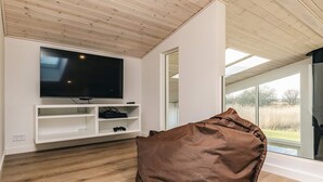 Ein modernes Wohnzimmer mit einem großen Flachbildfernseher an einer weißen Wand, Holzboden und einem großen Fenster mit Blick ins Freie.