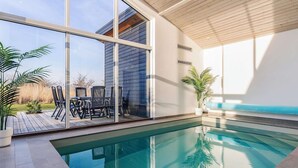 Ein moderner, überdachter Schwimmbereich mit großen Glaswänden, Holzbalkendecke und einer Terrassenanlage mit Stühlen und Pflanzen in Töpfen.