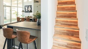 Ein modernes Interieur mit einer Holztreppe, einem Essbereich mit Stühlen und einem Tisch sowie großen Fenstern.