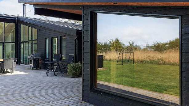 Eine moderne Terrasse im Freien mit Grill und Sitzgelegenheit, großen Fenstern und Blick auf eine Wiese.