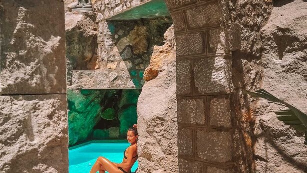 Un arc en pierre mène à un espace piscine où une personne est assise au bord de la piscine.