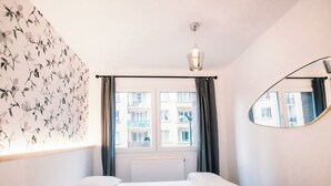 A bedroom with a bed, a window with curtains, a radiator, a wall-mounted mirror, and a floral wallpaper design. - Le Majestic - T2 en centre ville (Annecy)