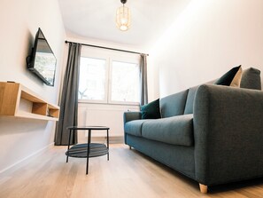 Un salon moderne avec un canapé gris, une petite table d’appoint noire, un téléviseur fixé au mur et une fenêtre avec des rideaux.