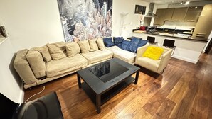 En moderne stue med sofa, sofabord og stol. I baggrunden ses et køkken med komfur og skabe.