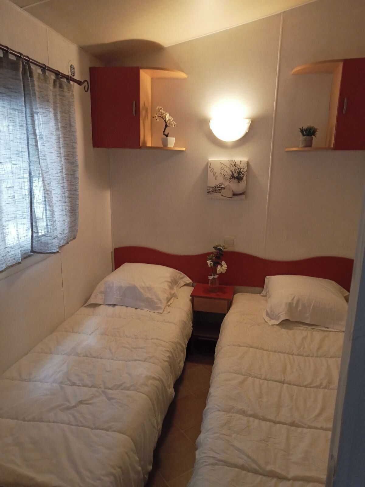 Un dormitorio con dos camas, una mesita de noche, una repisa en la pared con un jarrón y una lámpara de pared.