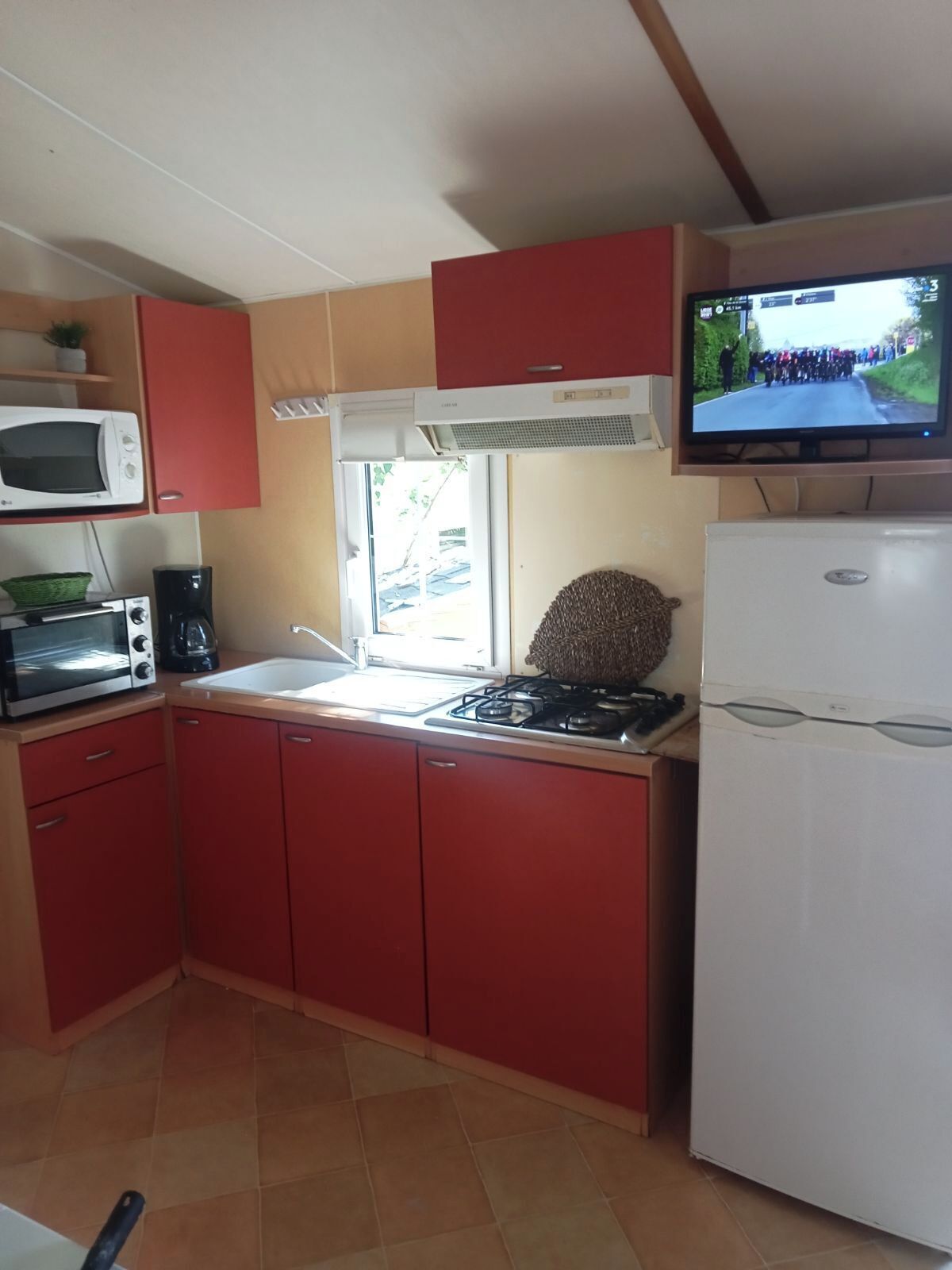 A compact kitchen with red cabinets, a white refrigerator, a stove, a microwave, and a television mounted above the stove.