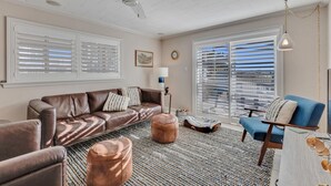 Un salon avec un canapé en cuir marron, un fauteuil bleu et une table basse. On y trouve un ventilateur de plafond, un lampadaire et une grande fenêtre avec des stores.