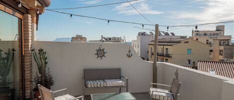 Ein Balkon mit einer Couch, zwei Stühlen und einem kleinen Tisch. Es sind Kübelpflanzen und ein dekoratives Wandelement zu sehen.