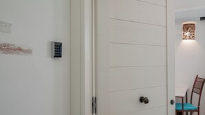 Une porte blanche dotée d’un clavier pour l’ouverture, une chaise en bois avec un coussin bleu et un luminaire fixé au mur.