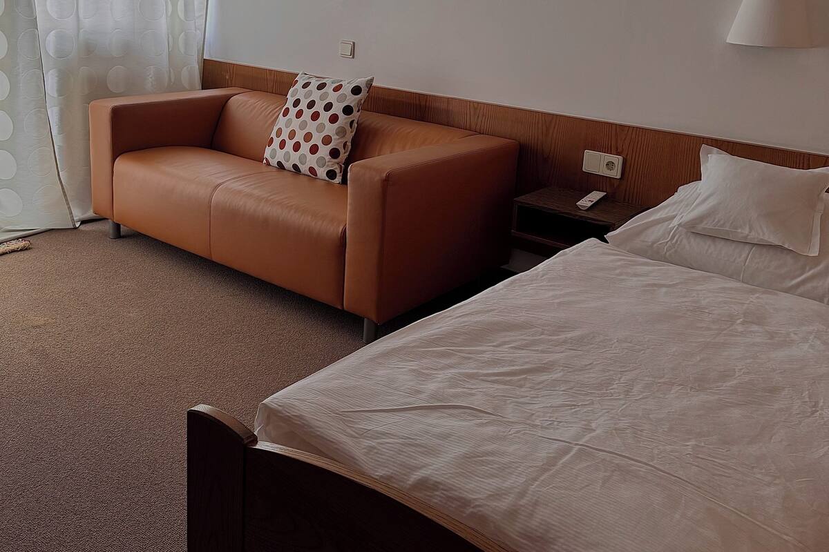 A hotel room with a brown leather sofa, a white bed with a white pillow, and a nightstand with a remote control.