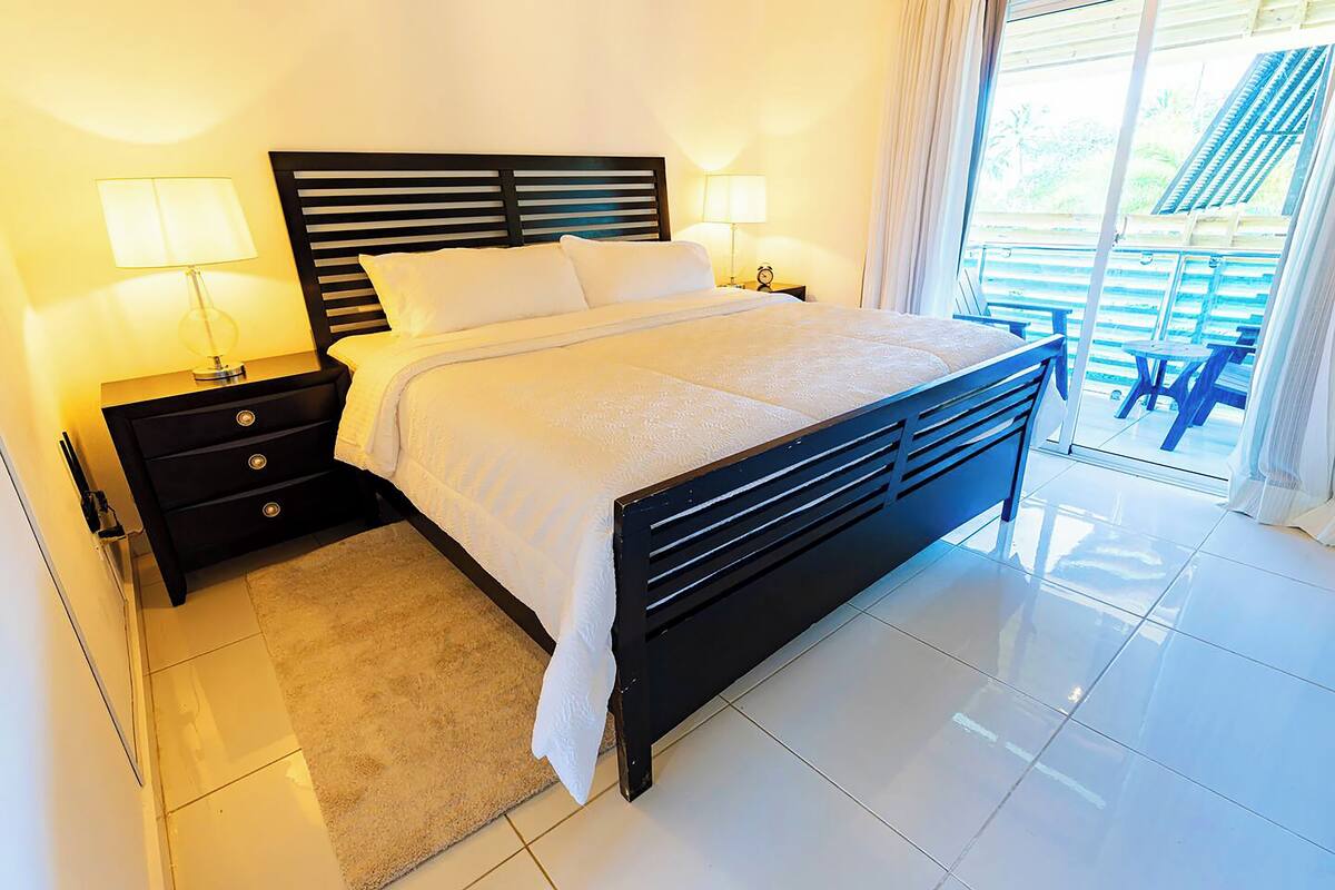 Un dormitorio moderno con una cama grande, mesitas de noche con lámparas y vistas a un balcón con mobiliario exterior.