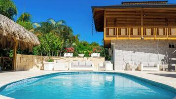 Relax by the serene pool surrounded by lush greenery and a traditional building with a thatched roof.