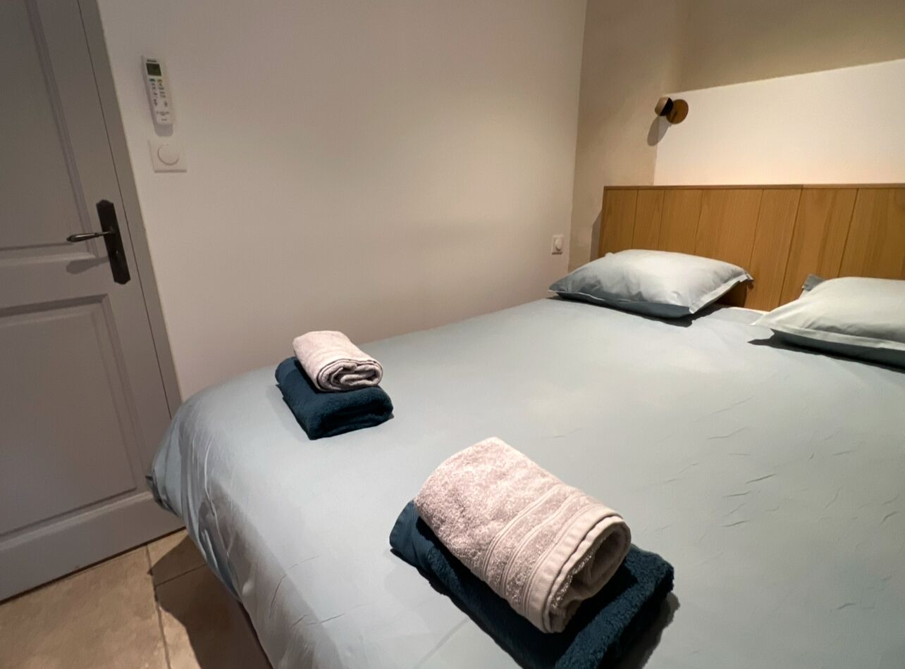 A hotel room with a bed, two pillows, a bedside table, a wall-mounted control panel, and a door.