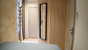 En smal hall med en säng längst in, en vägg klädd med träribbor och en dörr som leder till ett annat rum.