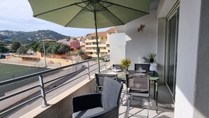 Ein Balkon mit Gartenmöbeln, einem Tisch und einem Sonnenschirm. Im Hintergrund ist ein Sportplatz und Gebäude zu sehen.