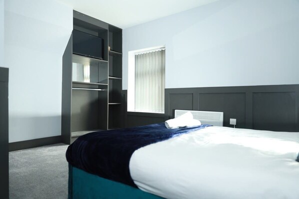 Une chambre d’hôtel avec un lit, une armoire intégrée, une fenêtre avec des stores et une tête de lit blanche.