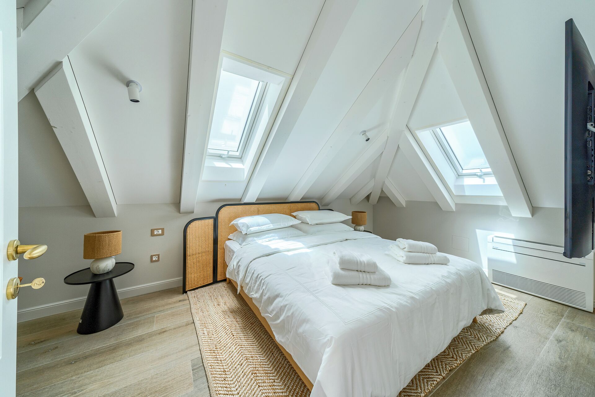 Un dormitorio con techo alto y tragaluces, una cama con ropa de cama blanca, una pequeña mesa redonda con lámpara, y un televisor de pantalla plana grande.