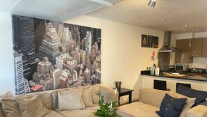A living room with a large cityscape painting, a sofa, a coffee table with a plant, and a kitchen area in the background.