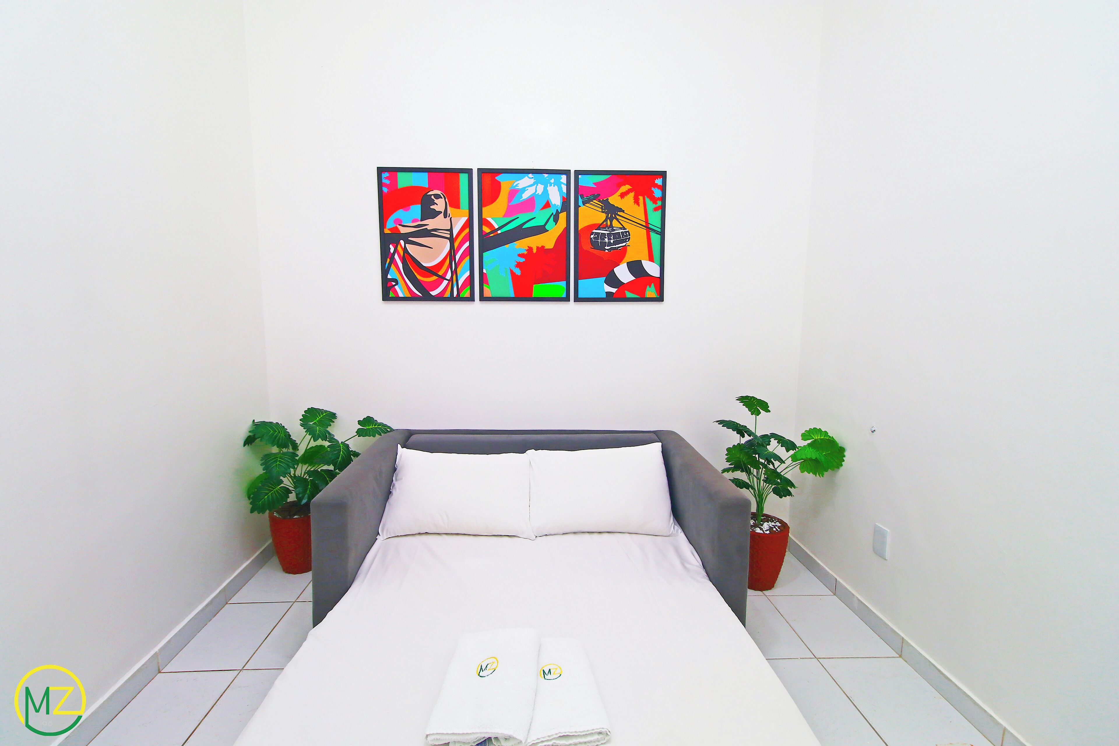 A hotel room with a bed, two potted plants, and three colorful paintings on the wall.