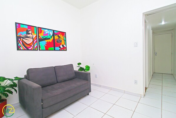 Een woonkamer met een grijze bank, een potplant en een kleurrijk abstract schilderij aan de muur.