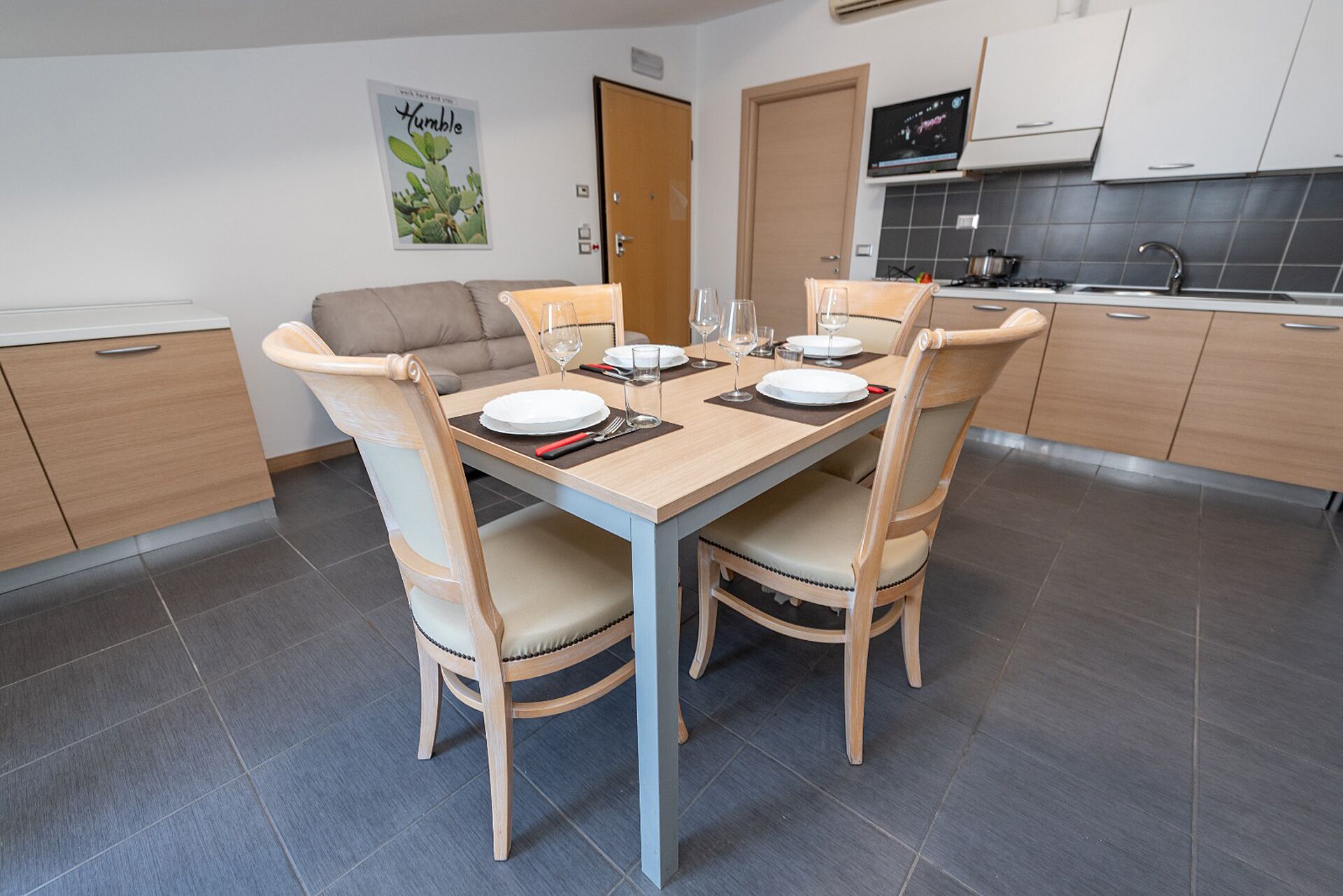 Una cocina moderna con una mesa de comedor para cuatro personas, armarios de madera clara y un suelo de baldosas grises.