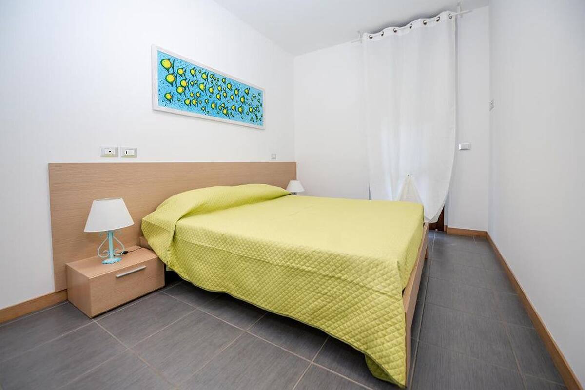 A neatly arranged hotel room with a bed, a bedside table with a lamp, a wall-mounted painting, and a window with a white curtain.