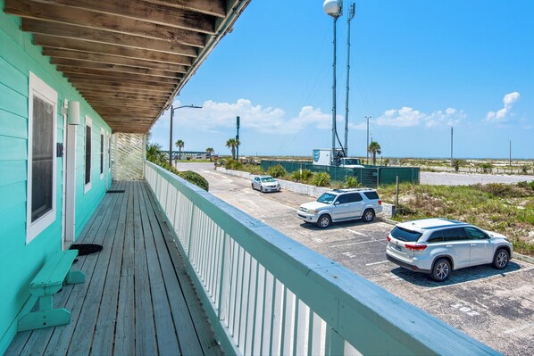 Un edificio turquesa con una terraza de madera que da vista a un estacionamiento y una playa.
