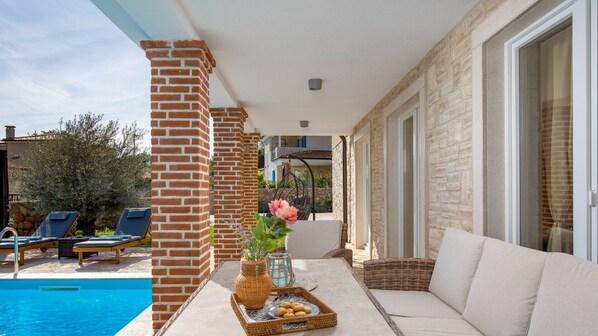 A poolside area with a brick column, a wicker chair, a cushioned sofa, and a table with a vase and snacks.