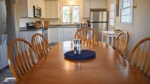 Un comedor con una mesa y sillas de madera, una cocina al fondo con armarios y electrodomésticos blancos, y una ventana que proporciona luz natural.