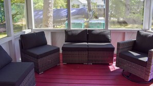 Un espacio al aire libre techado con mobiliario de mimbre y cojines, con vistas a un patio trasero con una casa y árboles.