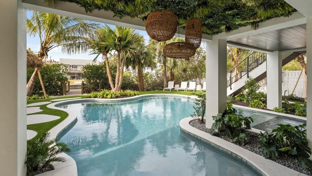 Una piscina con borde curvo, rodeada de jardines blancos y verdes, palmeras y cestas colgantes de mimbre.