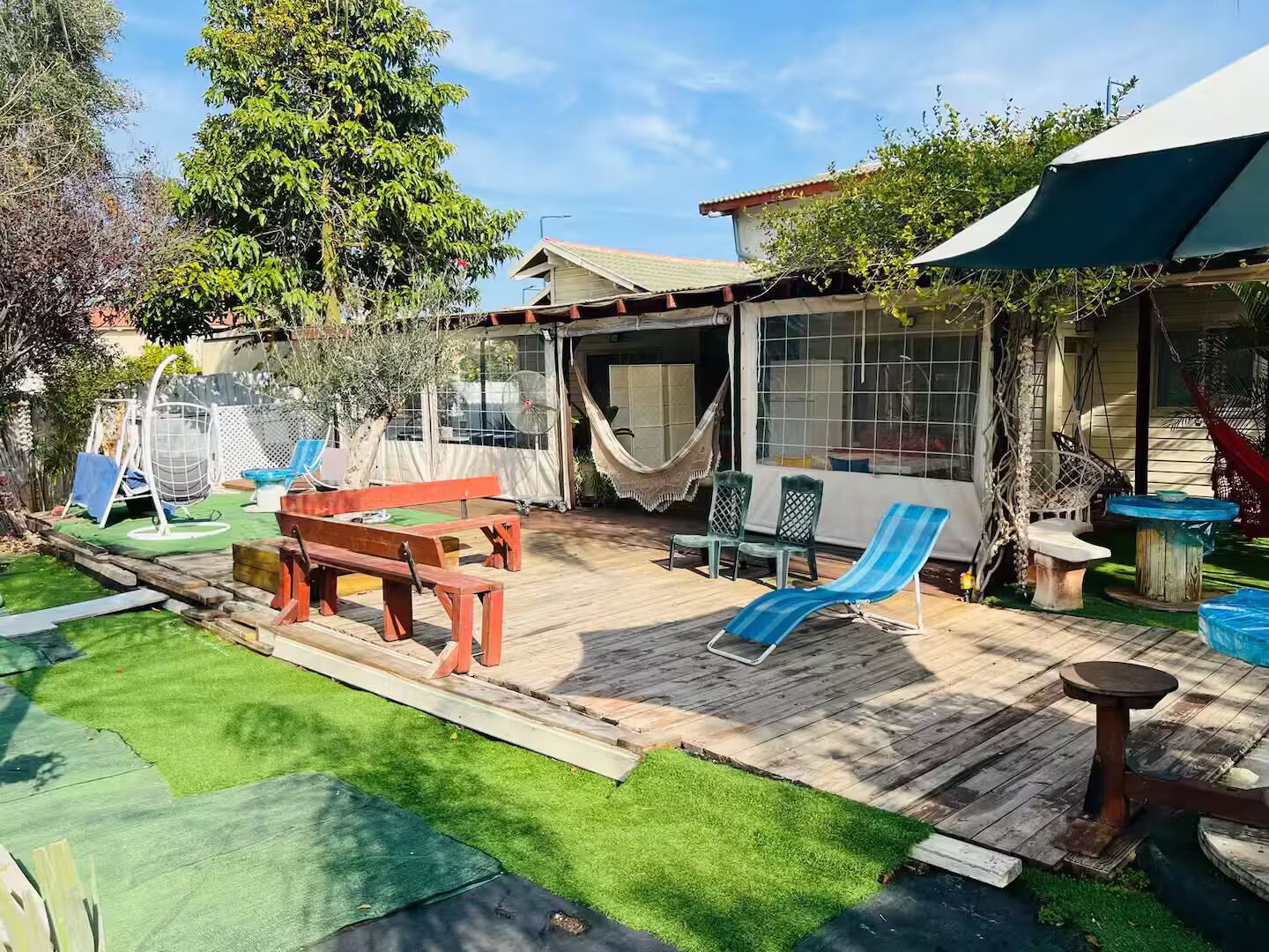 Un patio trasero con una terraza de madera, una hamaca, un conjunto de columpios y un área de patio techada.