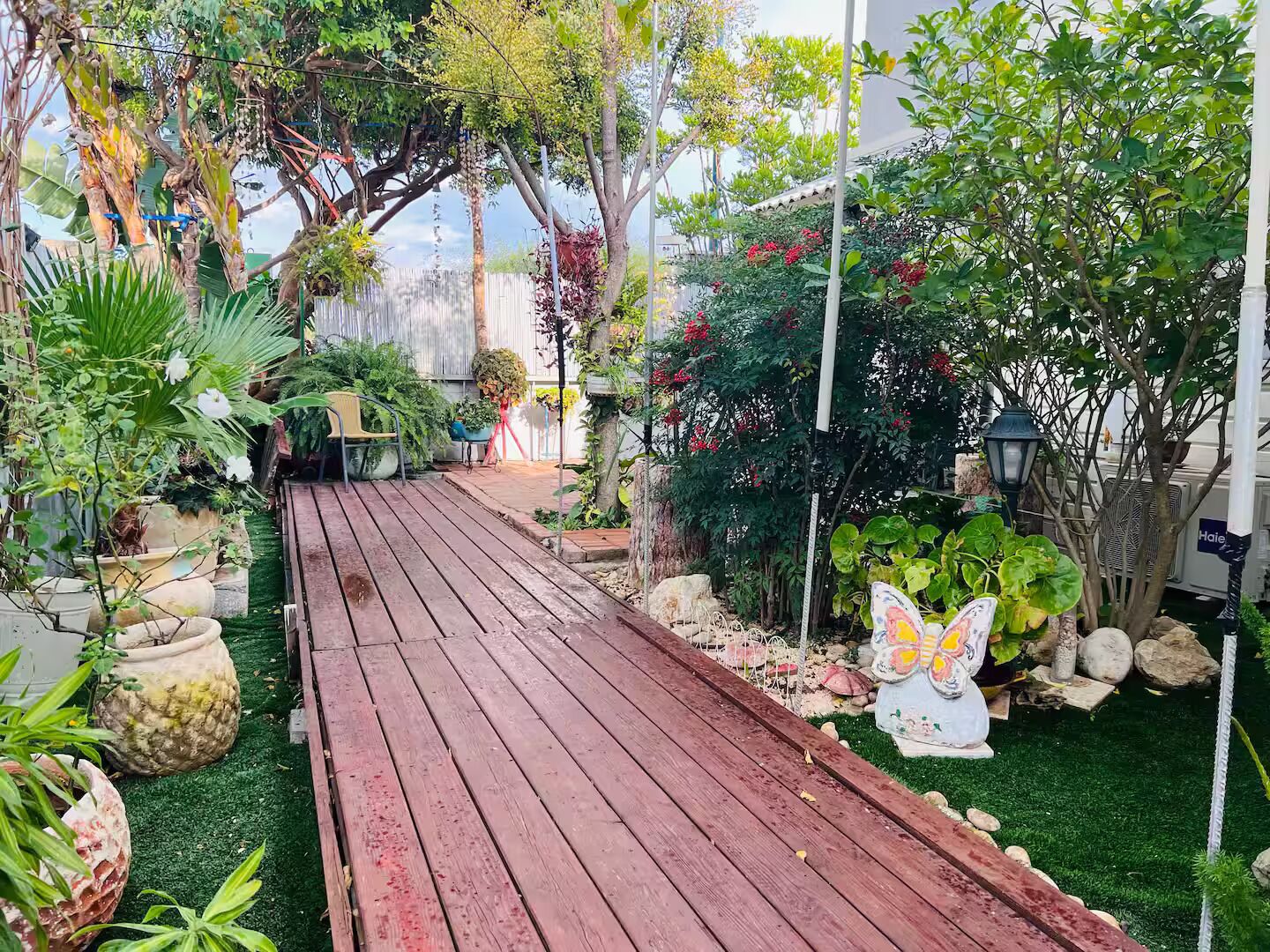 Una terraza de madera que conduce a un jardín con diversas plantas en macetas y una escultura de mariposa.