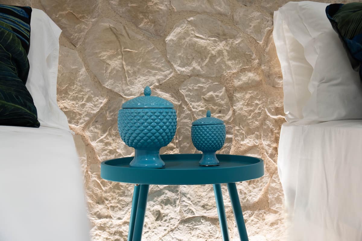A bed with white linens and two blue decorative items on a small round table against a textured stone wall.