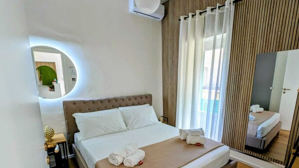 A hotel room with a bed, a round mirror, a bedside table with a lamp, and a window with curtains. - Lues Rooms (Ragusa)
