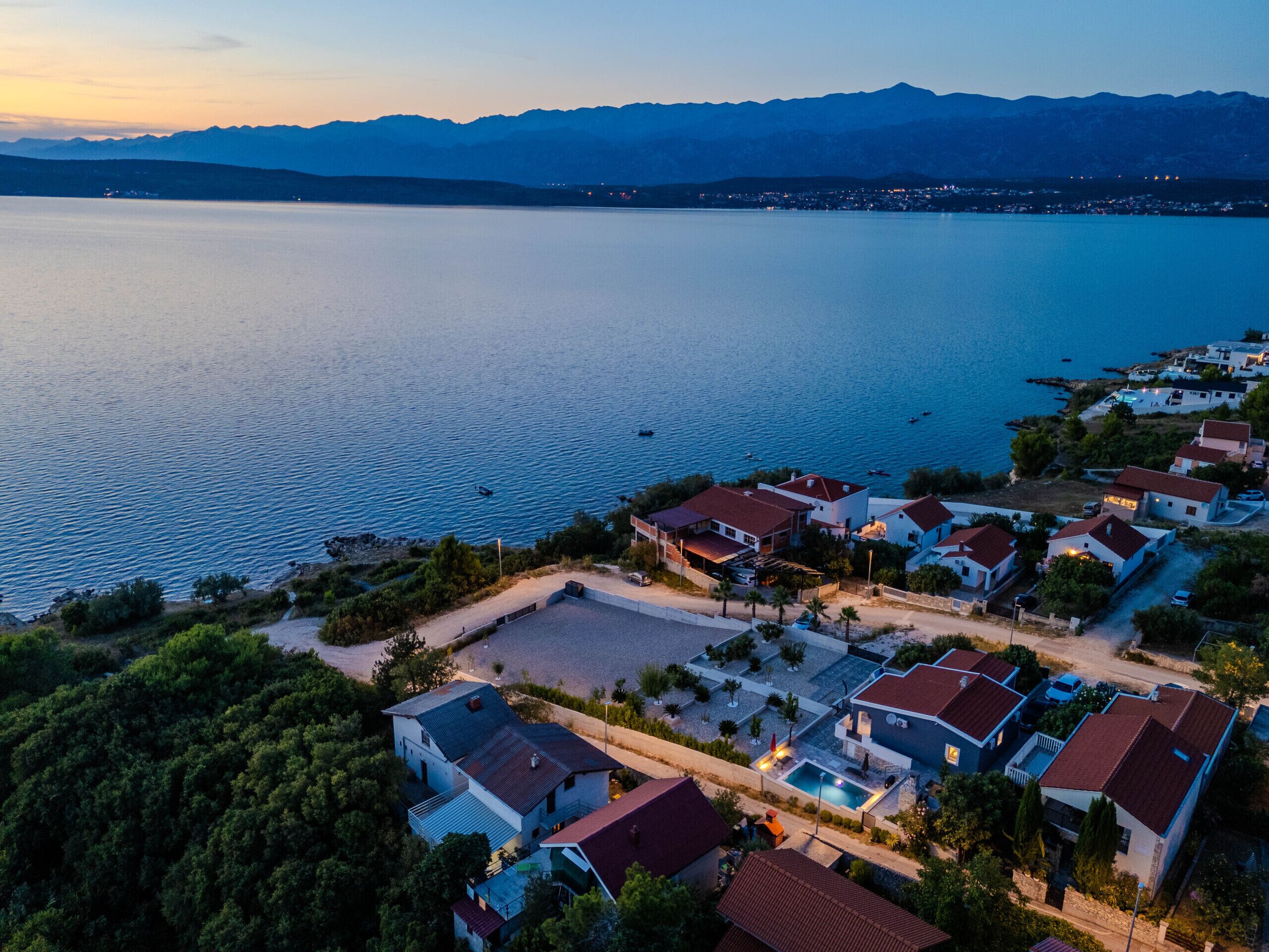 An aerial view of a coastal residential area with houses, a swimming pool, and a clear view of the sea and mountains in the background.