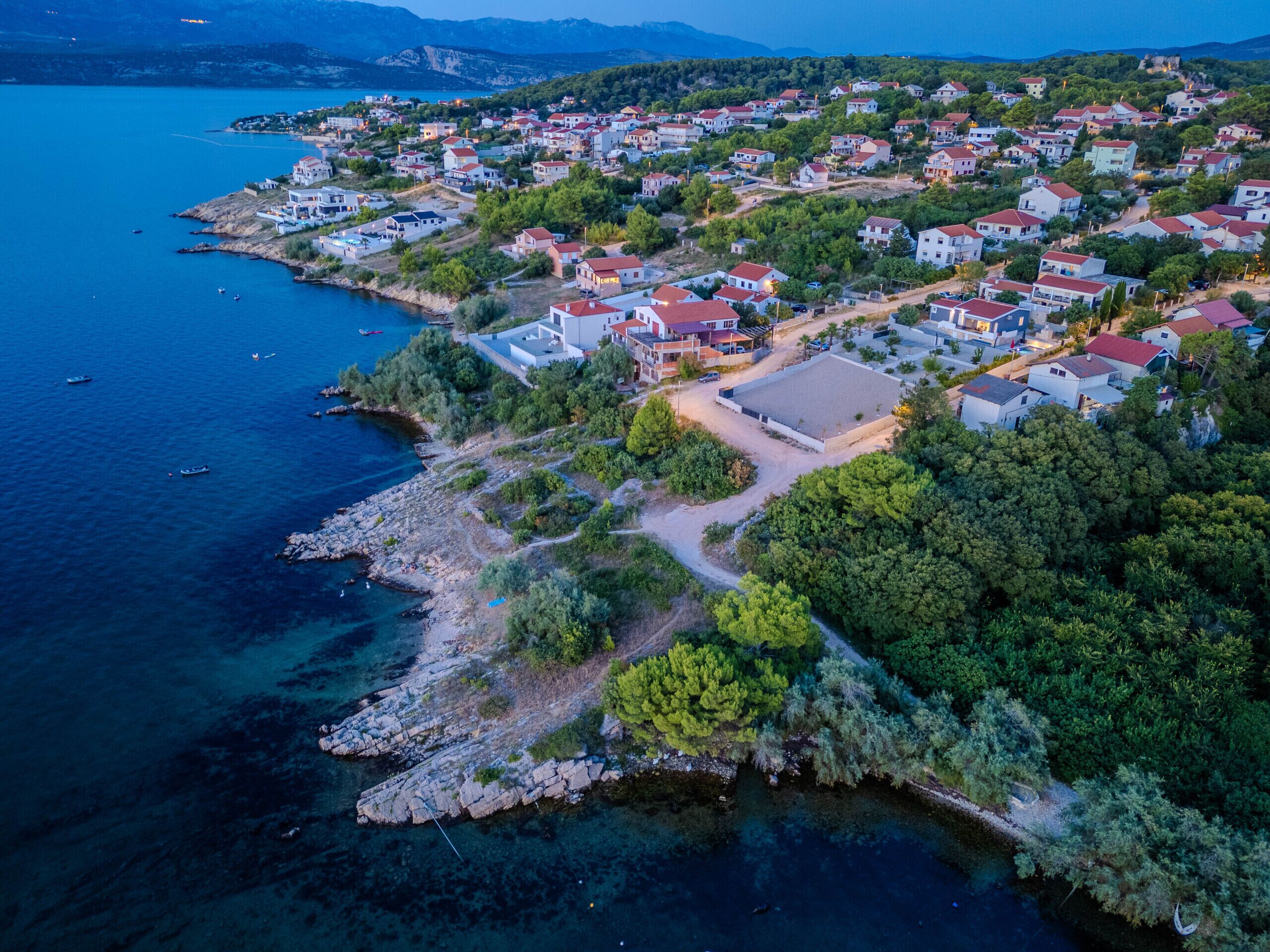 An aerial view of a coastal residential area with houses, a clear blue sea, and rocky terrain.