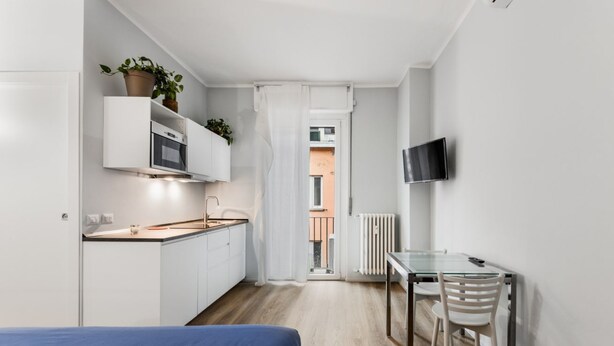 A compact kitchen with white cabinets, a microwave, and a potted plant. A glass table with a chair is positioned near a white wall with a mounted TV. - MI Life 23 APT -wifi-Fiera (Milano)