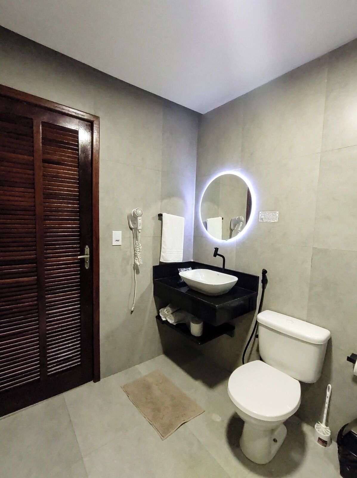 Un baño con puerta de madera, inodoro blanco, lavabo negro y espejo encima, además de un perchero para toallas.