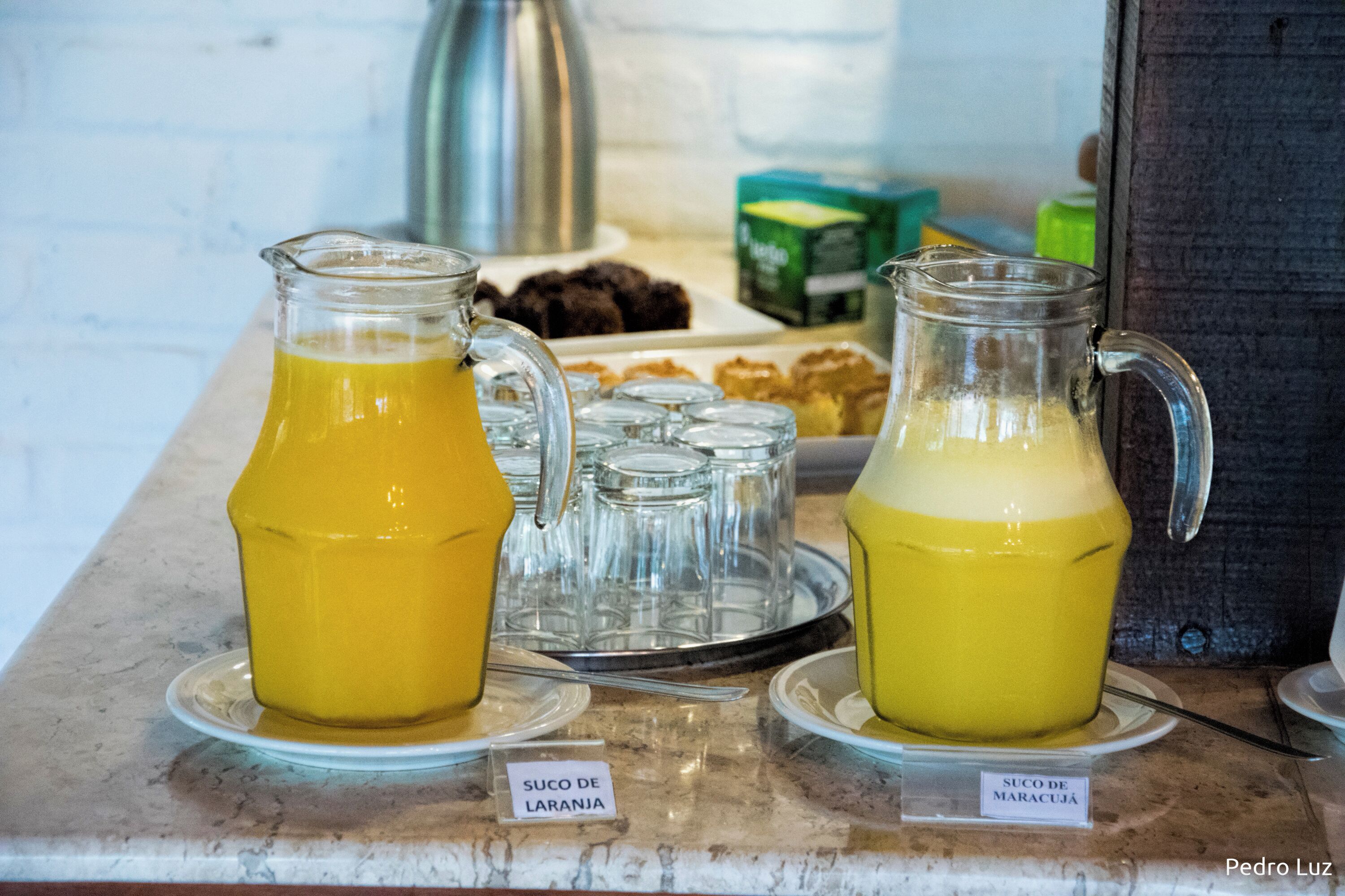 Um buffet de café da manhã com dois jarras de sucos de frutas, identificadas como 'Suco de Laranja' e 'Suco de Maracujá', em uma bancada de mármore.