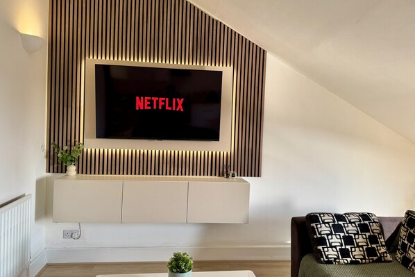 Un salon moderne avec un téléviseur fixé au mur qui affiche le mot « NETFLIX » en lettres rouges, un meuble de rangement blanc en dessous et un canapé agrémenté de coussins à motifs.