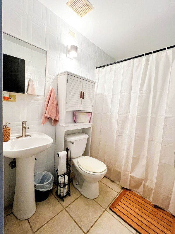 Un bagno con lavandino, water e mobiletti bianchi, una tenda da doccia e un tappetino di legno.