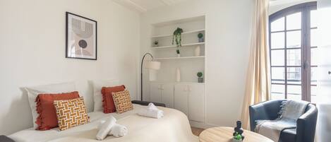 A modern hotel room with a bed, a small table, a chair, a shelf with plants, and a window with curtains.