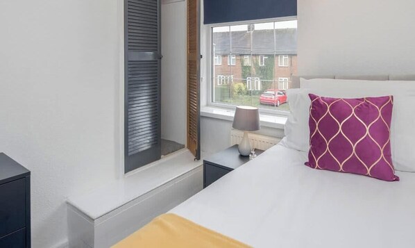 A bedroom with a bed, a nightstand, a lamp, a window with blinds, and a view of a red car and buildings outside.