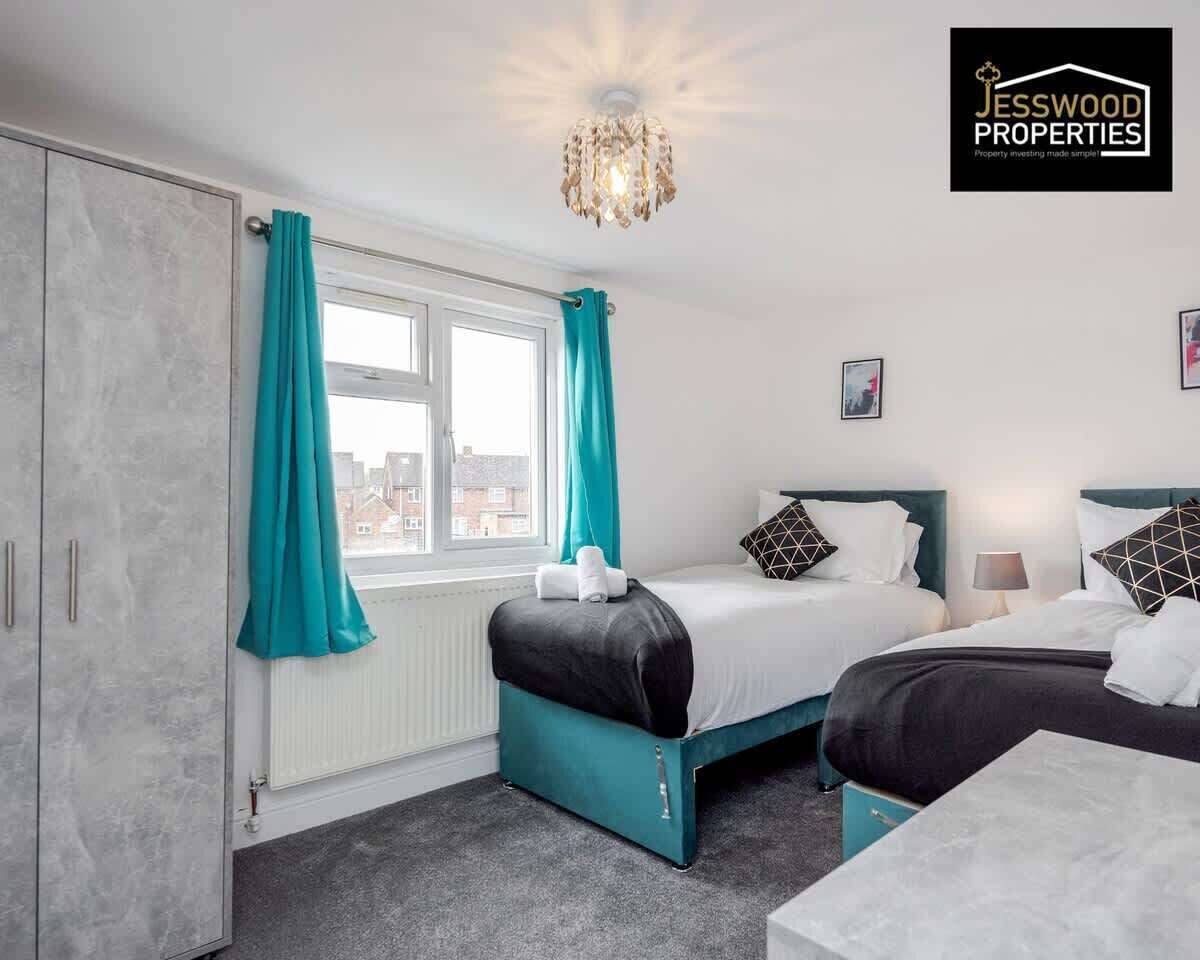 A bedroom with a bed, a chest of drawers, a window with curtains, a radiator, and a chandelier.