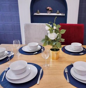 Ein Esstisch ist mit weißen Tellern, blauen Servietten und klaren Gläsern gedeckt, in der Mitte steht eine Vase mit weißen Blumen.